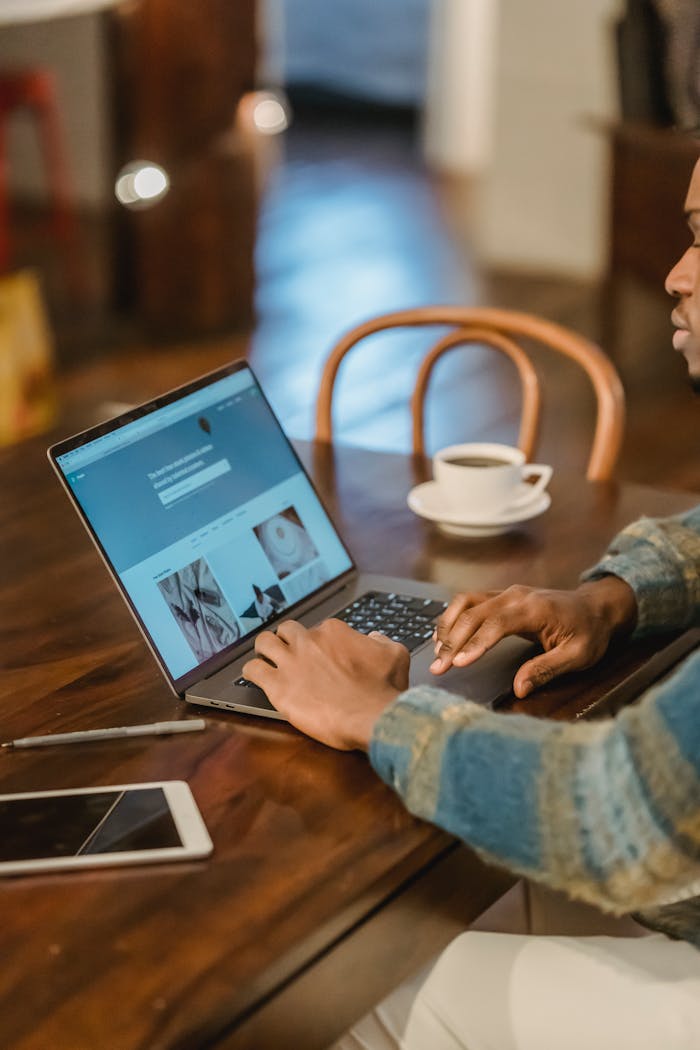 Crop unrecognizable Black male freelancer in casual clothes sitting at wooden table with laptop and typing on keyboard of modern interior