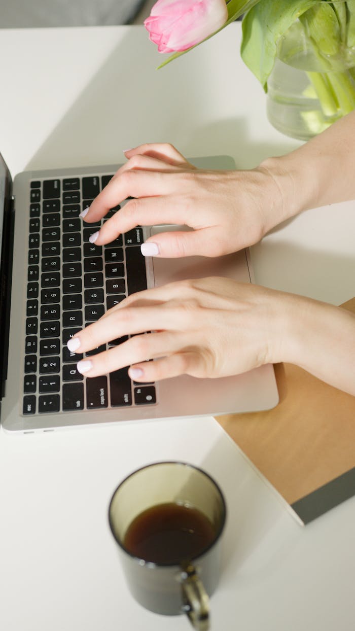 Hands typing on a laptop, with a pink tulip and coffee nearby on a desk